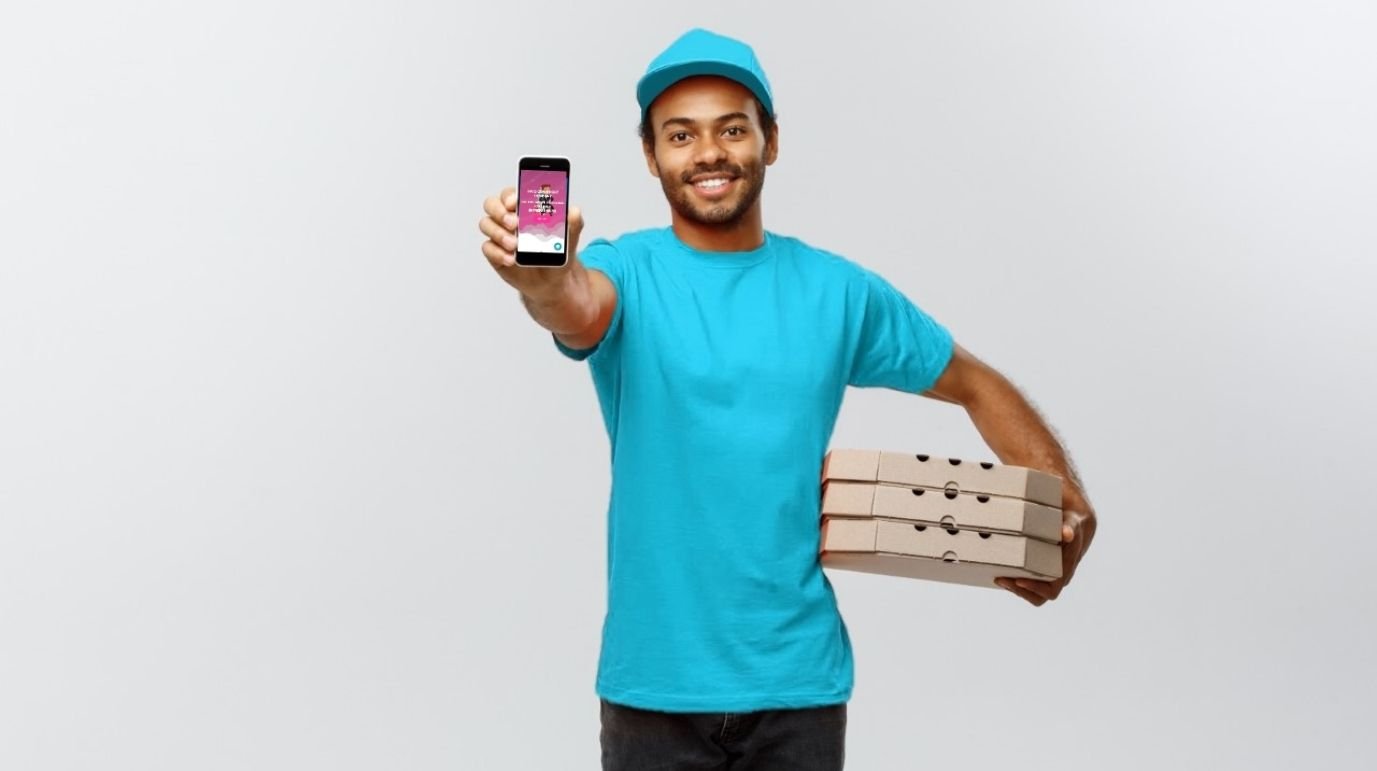Hymyilevä pizzalähetti turkoosissa asussa, pitelee puhelinta ja kantaa pizzalaatikoita